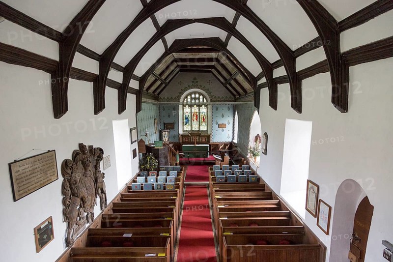 Raised angle view looking east down the nave towards the altar and east window with wooden pews, simple wood roof beams, whitewashed walls, interior of village parish church at Friston, Suffolk, England, UK. Mounted on the wall is a large Stuart Royal Coat of Arms of King James 1st 1605