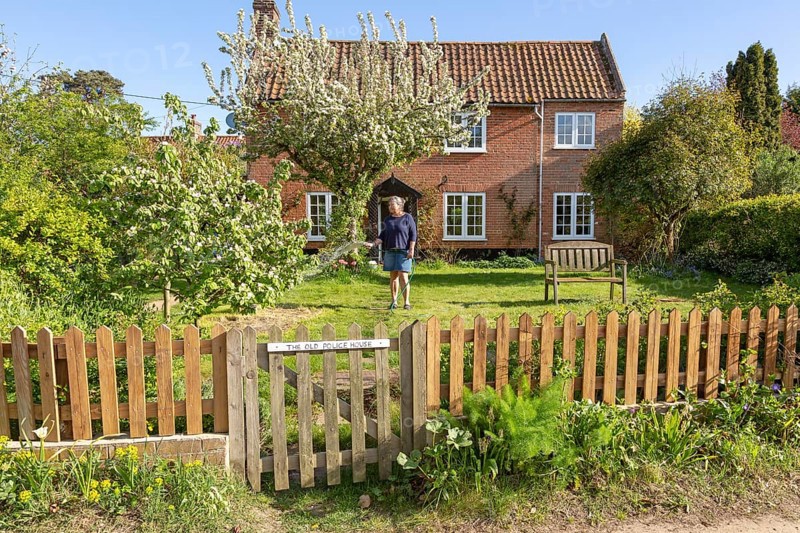 Woman watering garden with hosepipe in front of red brick historic detached house, Shottisham, Suffolk, England, UK