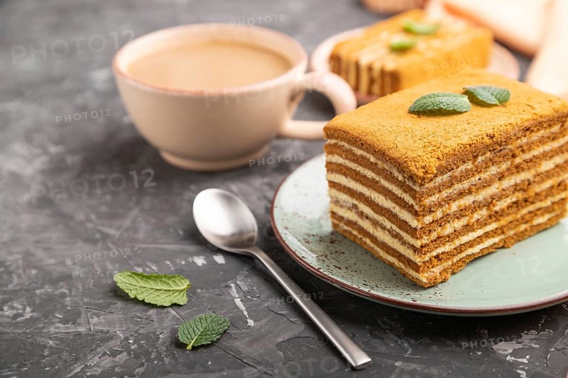 Homemade honey cake with milk cream and mint with cup of coffee on a black concrete background and orange textile. Side view, close up, selective focus