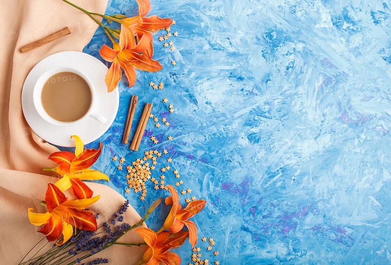 Orange day-lily and lavender flowers and a cup of coffee on a blue concrete background, with orange textile. Morninig, spring, fashion composition. Flat lay, top view, copy space