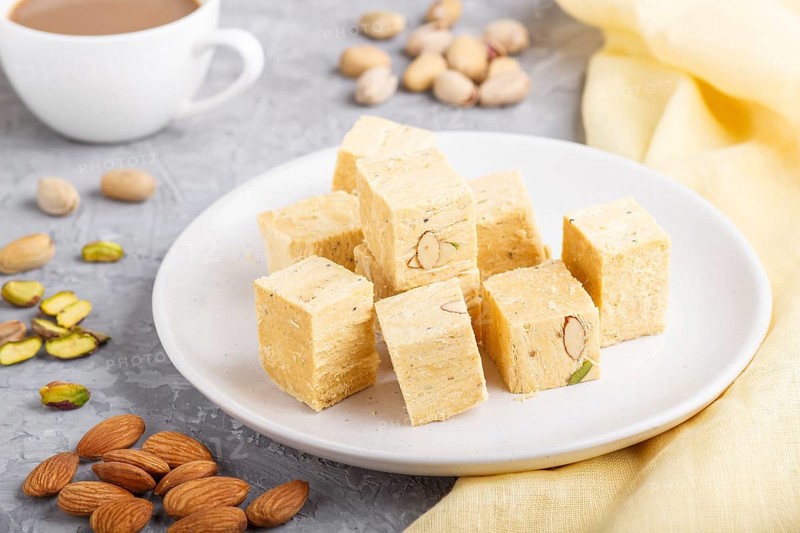 Traditional indian candy soan papdi in white plate with almond, pistache and a cup of coffee on a gray concrete background with yellow textile. side view, close up, selective focus