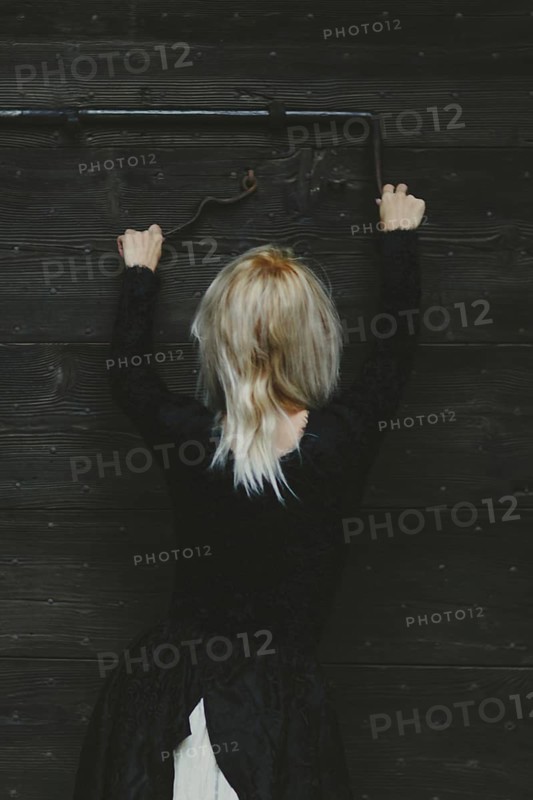 Back view of a blonde thin white woman dressed in black reaching up to a rusty antique metal lock on a wooden door with both hands
