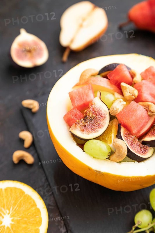 Vegetarian fruit salad of watermelon, grapes, figs, pear, orange, cashew on slate board on a black concrete background. Side view, close up, selective focus