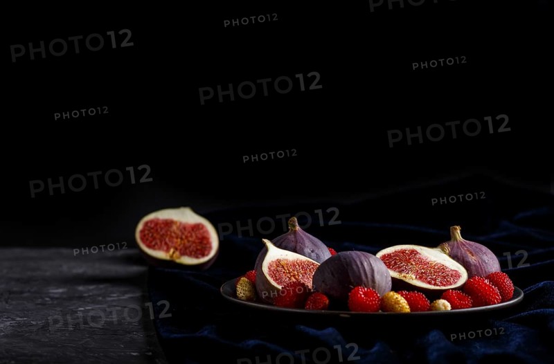 Fresh figs, strawberries and raspberries on blue ceramic plate on black concrete background and blue velvet textile. side view, close up, selective focus, low key