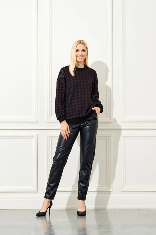 An elegant woman poses confidently against a white, panelled wall. She is dressed in a dark, patterned sweater and sleek black leather pants, complemented by classic black heels