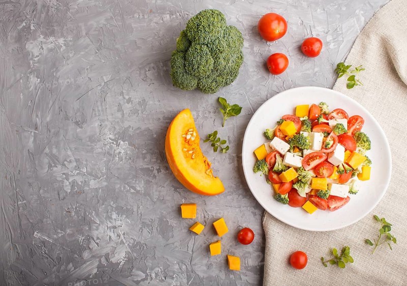 Vegetarian salad with broccoli, tomatoes, feta cheese, and pumpkin on white ceramic plate on a gray concrete background and linen textile, top view, copy space, flat lay