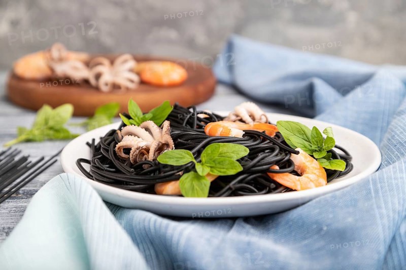 Black cuttlefish ink pasta with shrimps or prawns and small octopuses on gray wooden background and blue textile. Side view, close up, selective focus