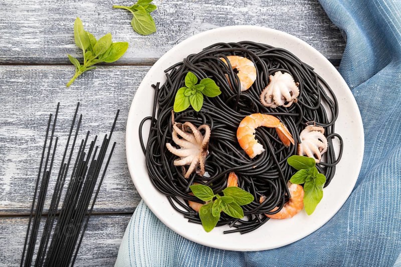 Black cuttlefish ink pasta with shrimps or prawns and small octopuses on gray wooden background and blue textile. Top view, flat lay, close up