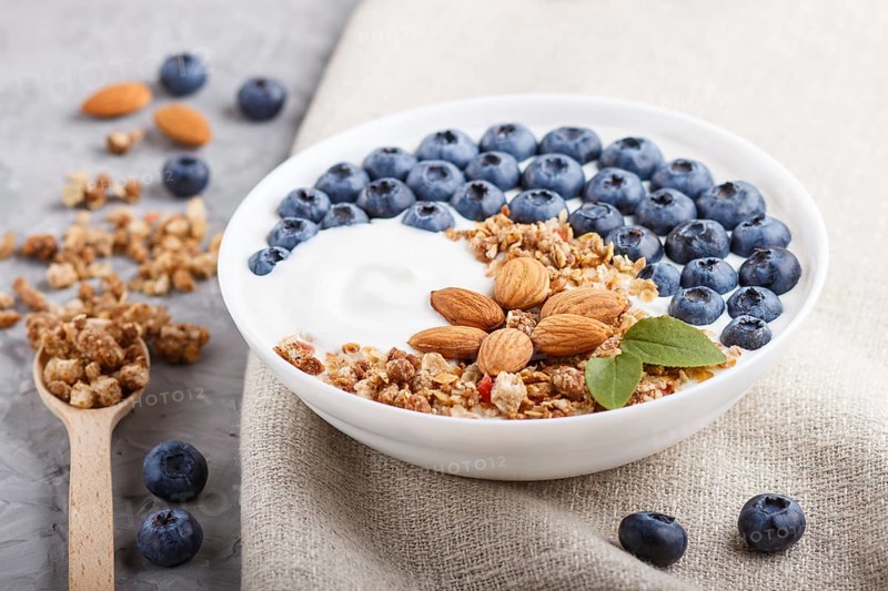Yoghurt with blueberry, granola and almond in white plate with wooden spoon on gray concrete background and linen textile. side view, close up, selective focus