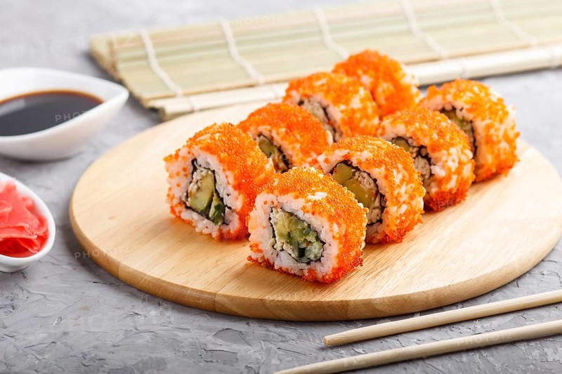 Japanese maki sushi rolls with flying fish roe, chopsticks, soy sauce and marinated ginger on wooden board on a gray concrete background. Side view, close up, selective focus