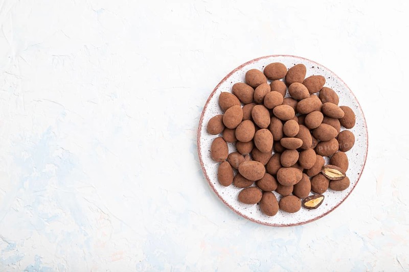 Almond in chocolate dragees on ceramic plate on white concrete background. Top view, copy space, flat lay
