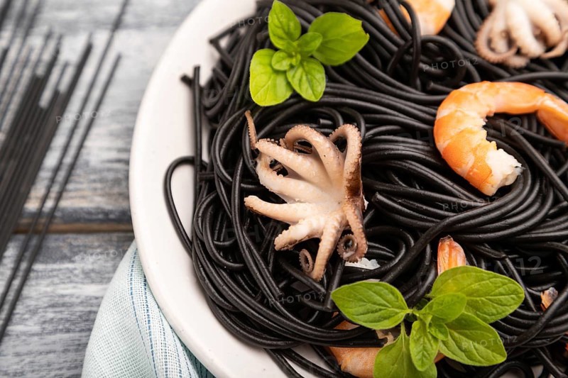 Black cuttlefish ink pasta with shrimps or prawns and small octopuses on gray wooden background and blue textile. Side view, close up, selective focus