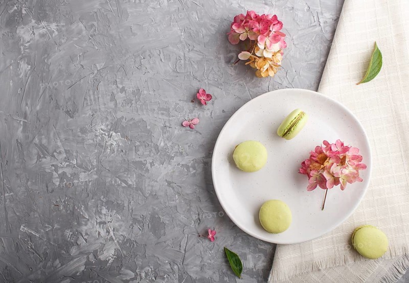 Green macarons or macaroons cakes on white ceramic plate on a gray concrete background and linen textile. Flat lay, top view, copy space