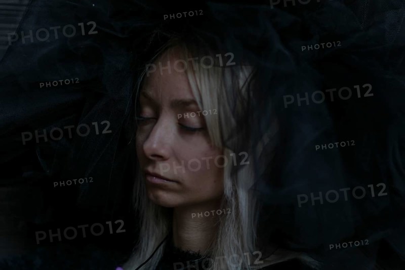 A young caucasian blonde woman's face is partially obscured by a dark veil, her eyes closed in a serene expression