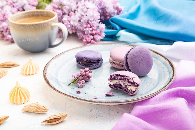 Purple macarons or macaroons cakes with cup of coffee on a white concrete background and magenta-blue textile. Side view, close up