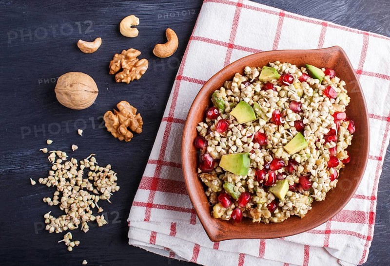 Salad of germinated buckwheat, avocado, walnut and pomegranate seeds in clay plate on black wooden background. Top view, flat lay