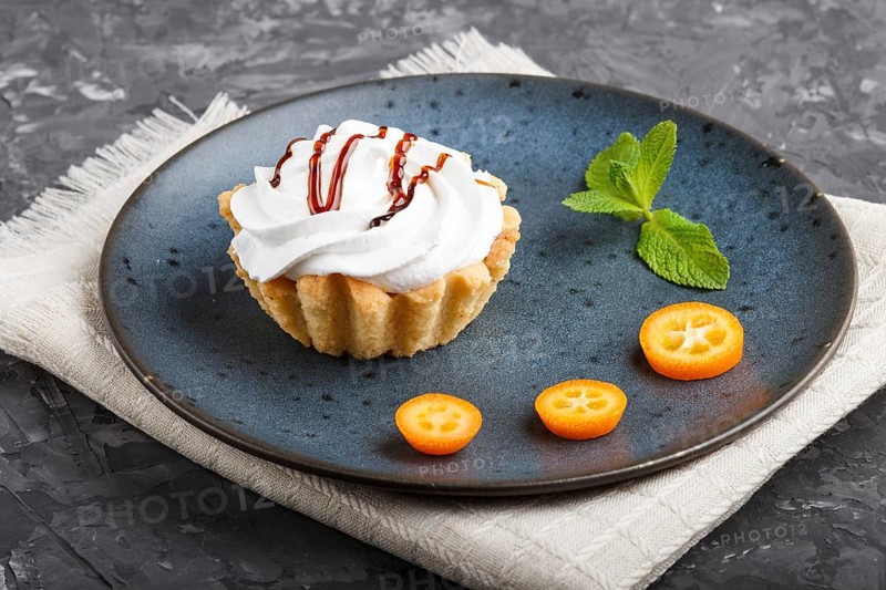Cake with whipped egg cream on a blue ceramic plate with kumquat slices and mint leaves on a black concrete background with linen napkin. side view