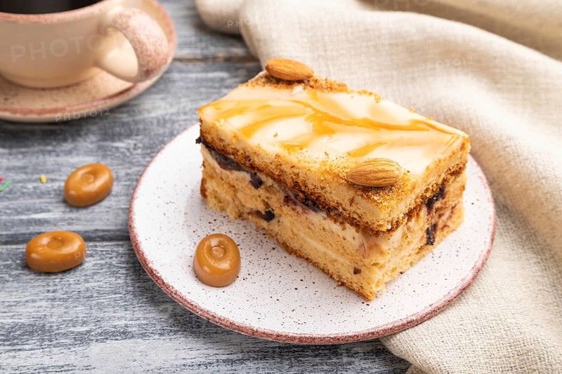 Honey cake with milk cream, caramel, almonds and a cup of coffee on a gray wooden background and linen textile. Side view, close up