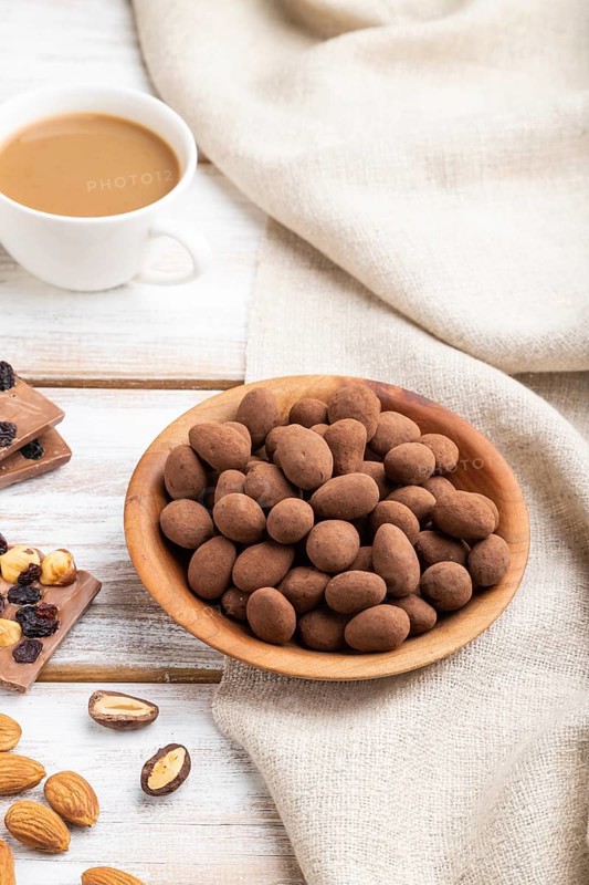 Almond in chocolate dragees in wooden plate and a cup of coffee on white wooden background and linen textile. Side view, close up