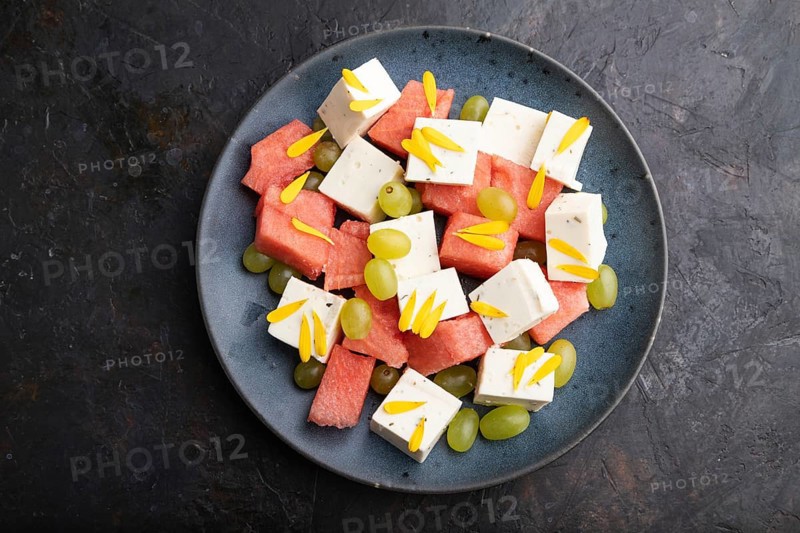 Vegetarian salad with watermelon, feta cheese, and grapes on blue ceramic plate on black concrete background. top view, close up, flat lay