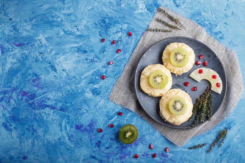 Pieces of baked pork with pineapple, cheese and kiwi on blue background, top view, copy space