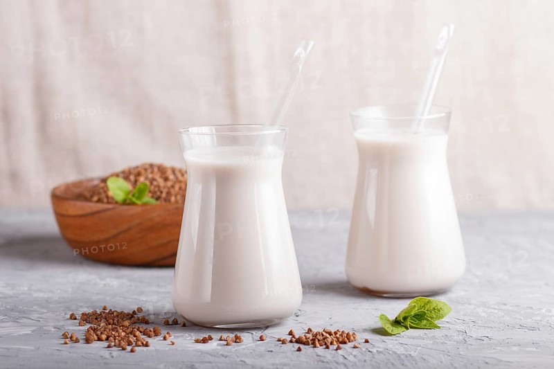 Organic non dairy buckwheat milk in glass and wooden plate with buckwheat seeds on a gray concrete background. Vegan healthy food concept, close up, side view