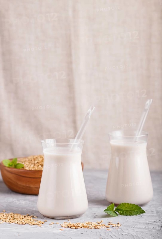 Organic non dairy oats milk in glass and wooden plate with oats seeds on a gray concrete background. Vegan healthy food concept, close up, side view, copy space