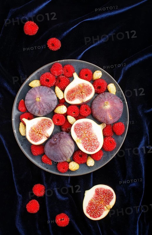 Fresh figs, strawberries and raspberries on blue ceramic plate on black concrete background and blue velvet textile. top view, flat lay, close up
