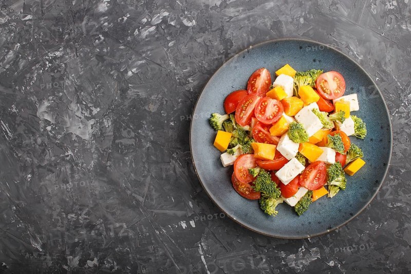 Vegetarian salad with broccoli, tomatoes, feta cheese, and pumpkin on a blue ceramic plate on a black concrete background, top view, copy space, flat lay