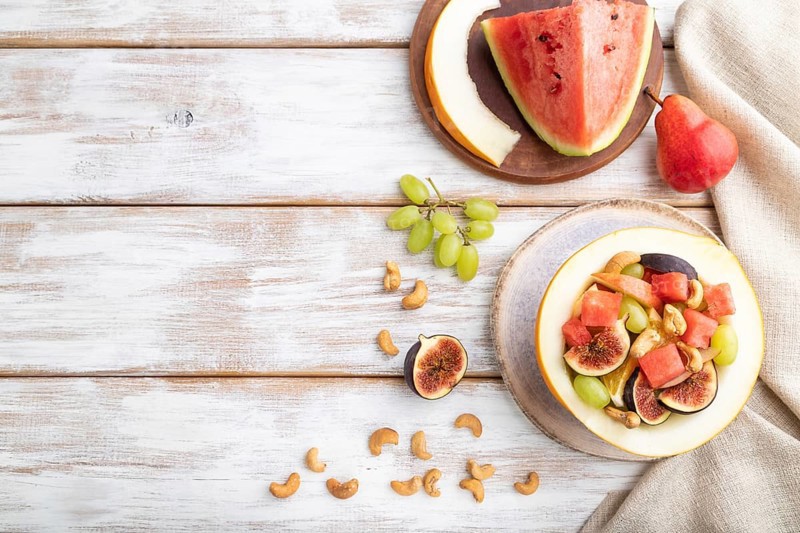 Vegetarian fruit salad of watermelon, grapes, figs, pear, orange, cashew on white wooden background and linen textile. Top view, flat lay, copy space
