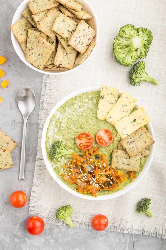 Green broccoli cream soup with crackers and cheese in white bowl on a gray concrete background and linen napkin. top view, flat lay, close up