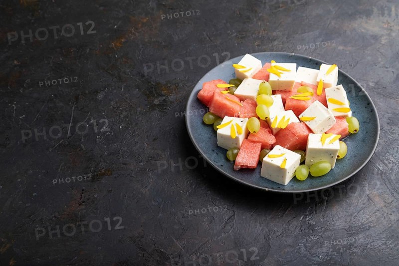 Vegetarian salad with watermelon, feta cheese, and grapes on blue ceramic plate on black concrete background. Side view, copy space