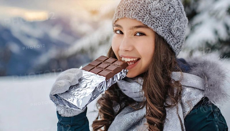KI generated, Young girl, 15, years, eating a bar of chocolate, one person, outdoor shot, ice, snow, winter, seasons, eating, eating, hat, bobble hat, gloves, winter jacket, cold, coldness