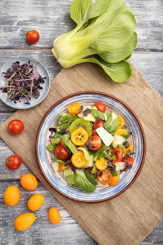 Vegetarian salad of pac choi cabbage, kiwi, tomatoes, kumquat, microgreen sprouts on gray wooden background and linen textile. Top view, flat lay, close up