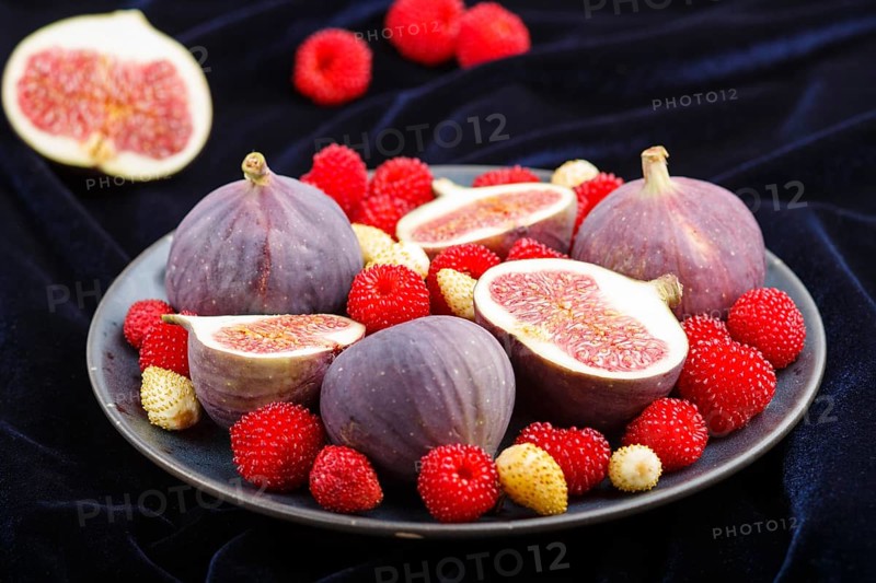 Fresh figs, strawberries and raspberries on blue ceramic plate on black concrete background and blue velvet textile. side view, close up, selective focus