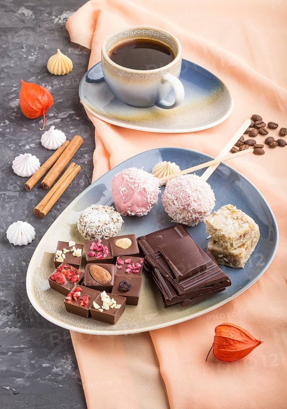 A pieces of homemade chocolate with coconut candies and a cup of coffee on a black concrete background and orange textile. side view, close up