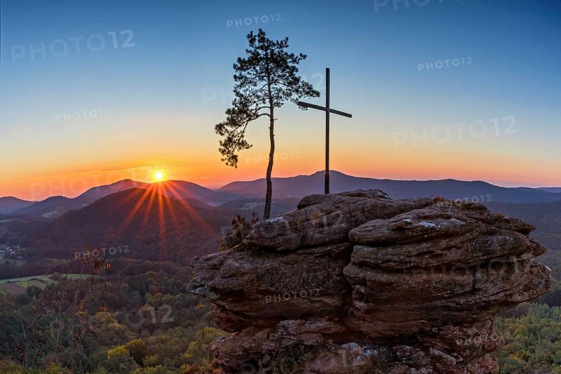 Sunrise on the Roetzenfelsen in the Palatinate Forest with bright sunshine and the lone pine tree and the summit cross on the red rock