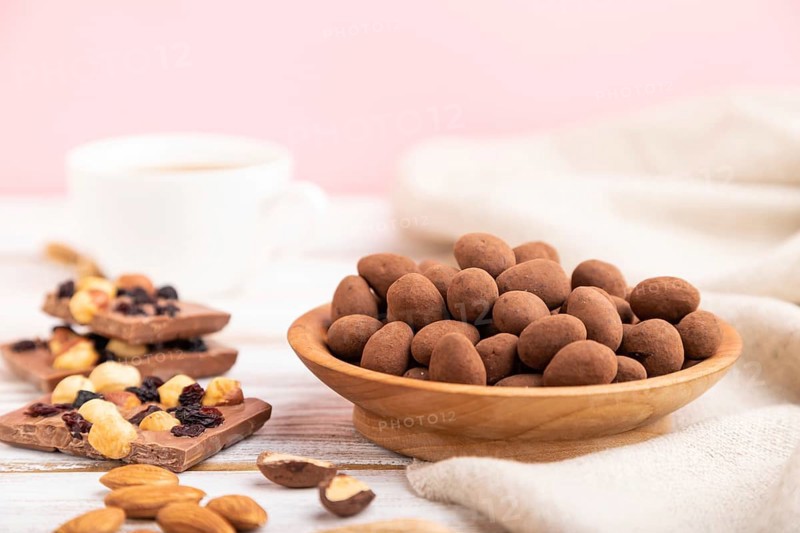 Almond in chocolate dragees in wooden plate and a cup of coffee on white and pink background and linen textile. Side view, copy space, selective focus