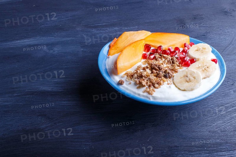 A blue plate with greek yogurt, granola, persimmon, banana, pomegranate black wooden background. close up. copy space