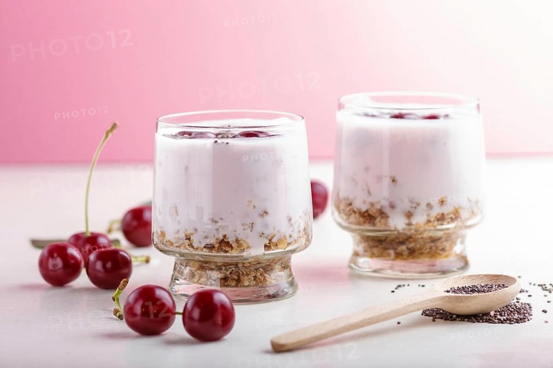 Yoghurt with cherries, chia seeds and granola in glass with wooden spoon on pink and white background. side view, close up, selective focus, copy space