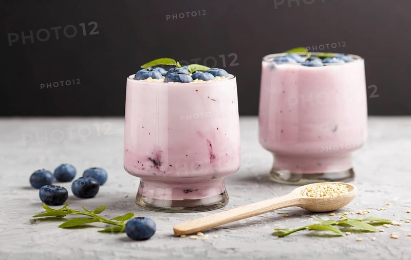 Yoghurt with blueberry and sesame in a glass and wooden spoon on gray and black background. side view, close up