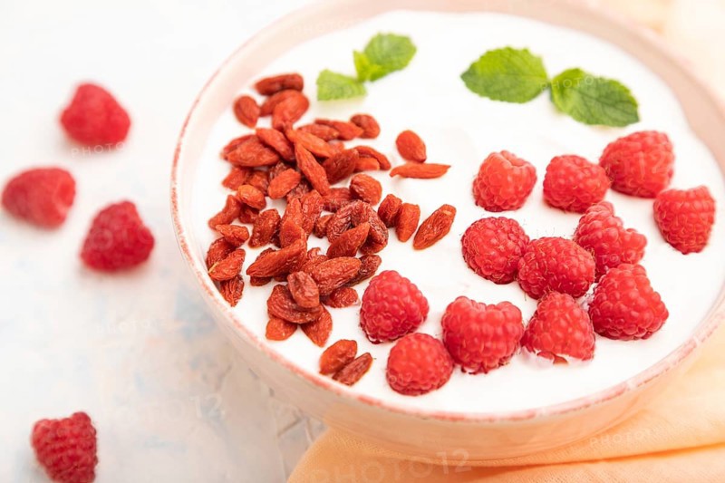 Yogurt with raspberry and goji berries in ceramic bowl on white concrete background and orange linen textile. Side view, close up, selective focus