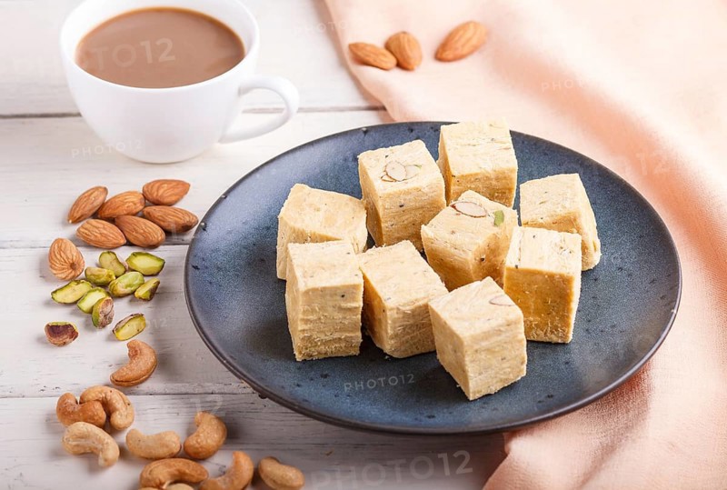 Traditional indian candy soan papdi in a blue ceramic plate with almond, pistache, cashew and a cup of coffee on a white wooden background with orange textile. side view, close up, selective focus