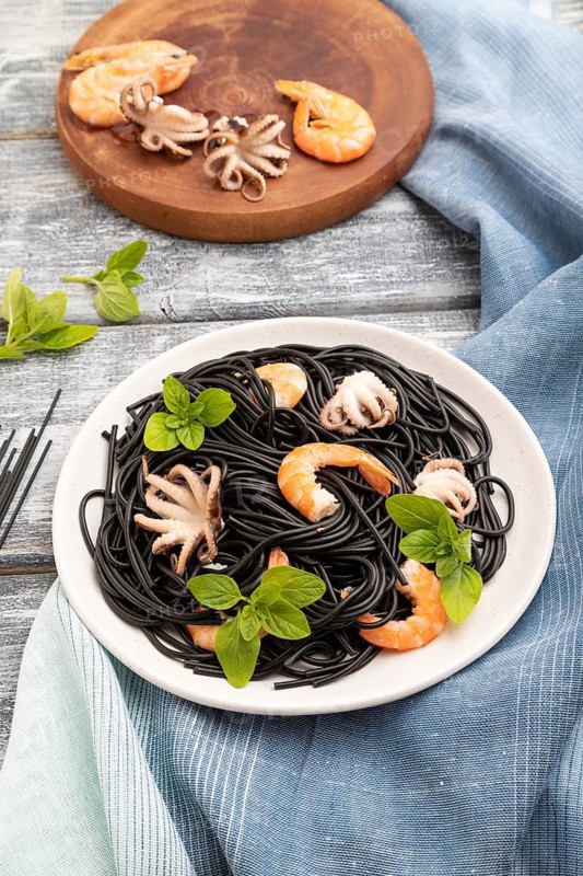 Black cuttlefish ink pasta with shrimps or prawns and small octopuses on gray wooden background and blue textile. Side view, close up