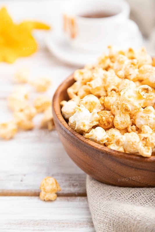 Popcorn with caramel in wooden bowl and a cup of coffee on a white wooden background and linen textile. Side view, close up, selective focus
