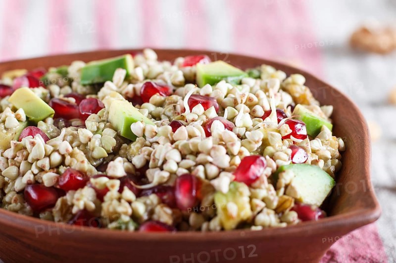 Salad of germinated buckwheat, avocado, walnut and pomegranate seeds in clay plate on white wooden background. Side view, close up, selective focus