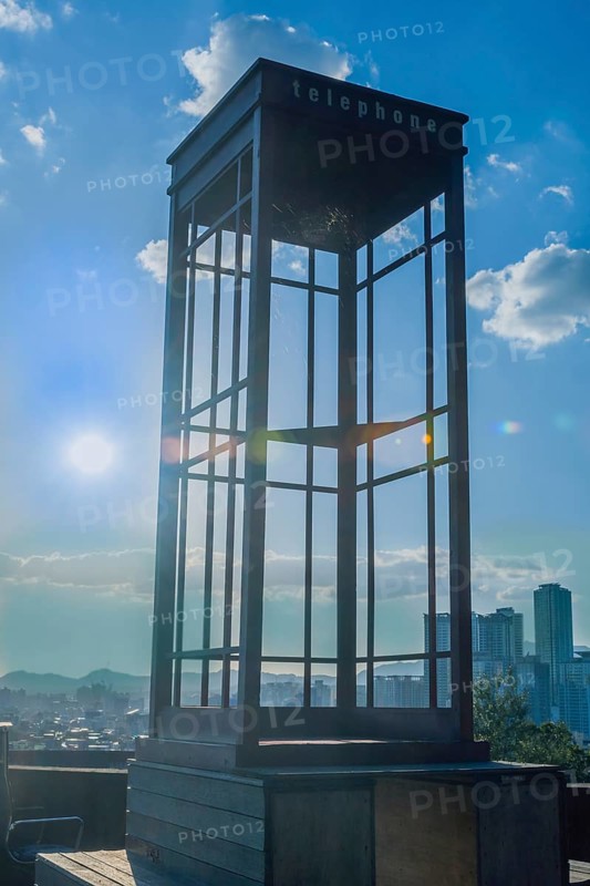 Red wooden telephone booth minus phone and without glass on roof of building back lit by the sun with blue sky and clouds in background in South Korea