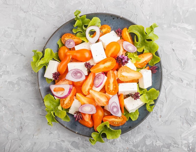 Vegetarian salad with fresh grape tomatoes, feta cheese, lettuce and onion on blue ceramic plate on gray concrete background, top view, close up, flat lay