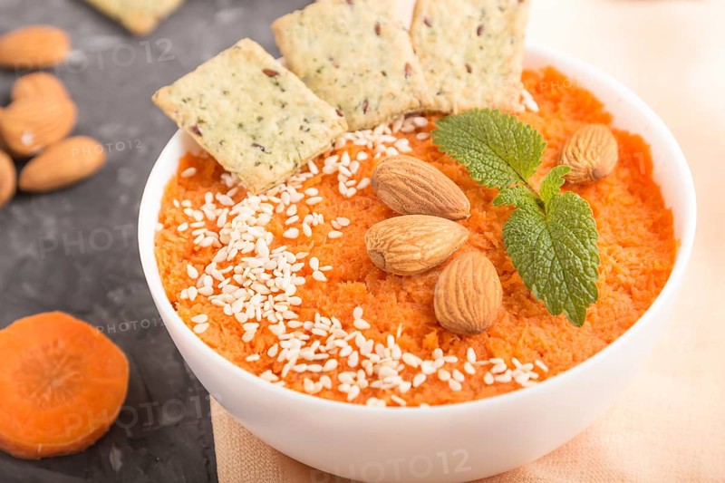 Carrot cream soup with sesame seeds, almonds and snacks in white bowl on a black concrete background with orange textile. side view, close up, selective focus