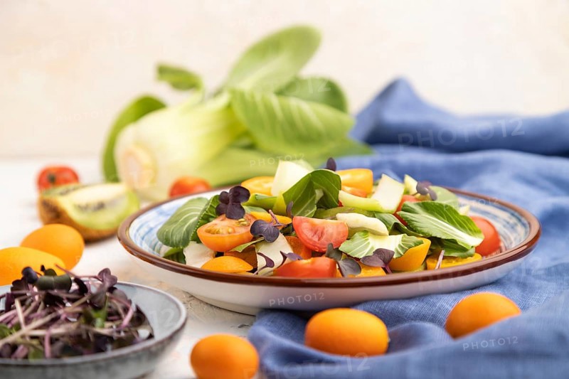 Vegetarian salad of pac choi cabbage, kiwi, tomatoes, kumquat, microgreen sprouts on a white concrete background and blue linen textile. Side view, close up, selective focus